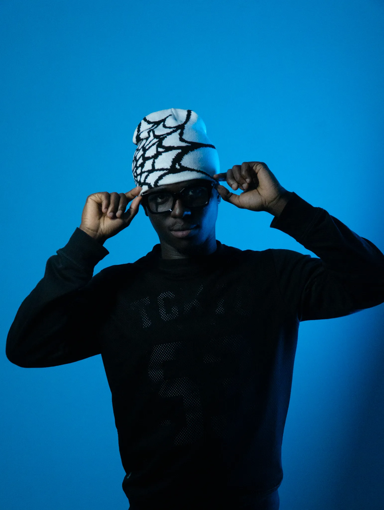 Studio fashion portrait of a model adjusting a graphic beanie set against a solid blue backdrop